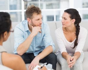 A couple, seated on a couch, is pondering a question from a counsellor. An example of couples counselling in Tantramar, representing an in-person session.